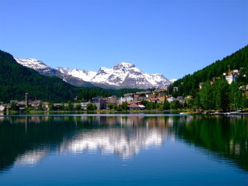 St. Moritz flanked by tree covered hills and a snow covered mountain in the background, reflected in a tranquil lake, Switzerland