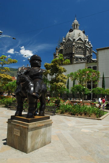 The artist Botero’s ‘Man on Horse Statue’ stands in front of bright trees and flowers in and an ornate building in Plaza Botero, Medellín, Colombia