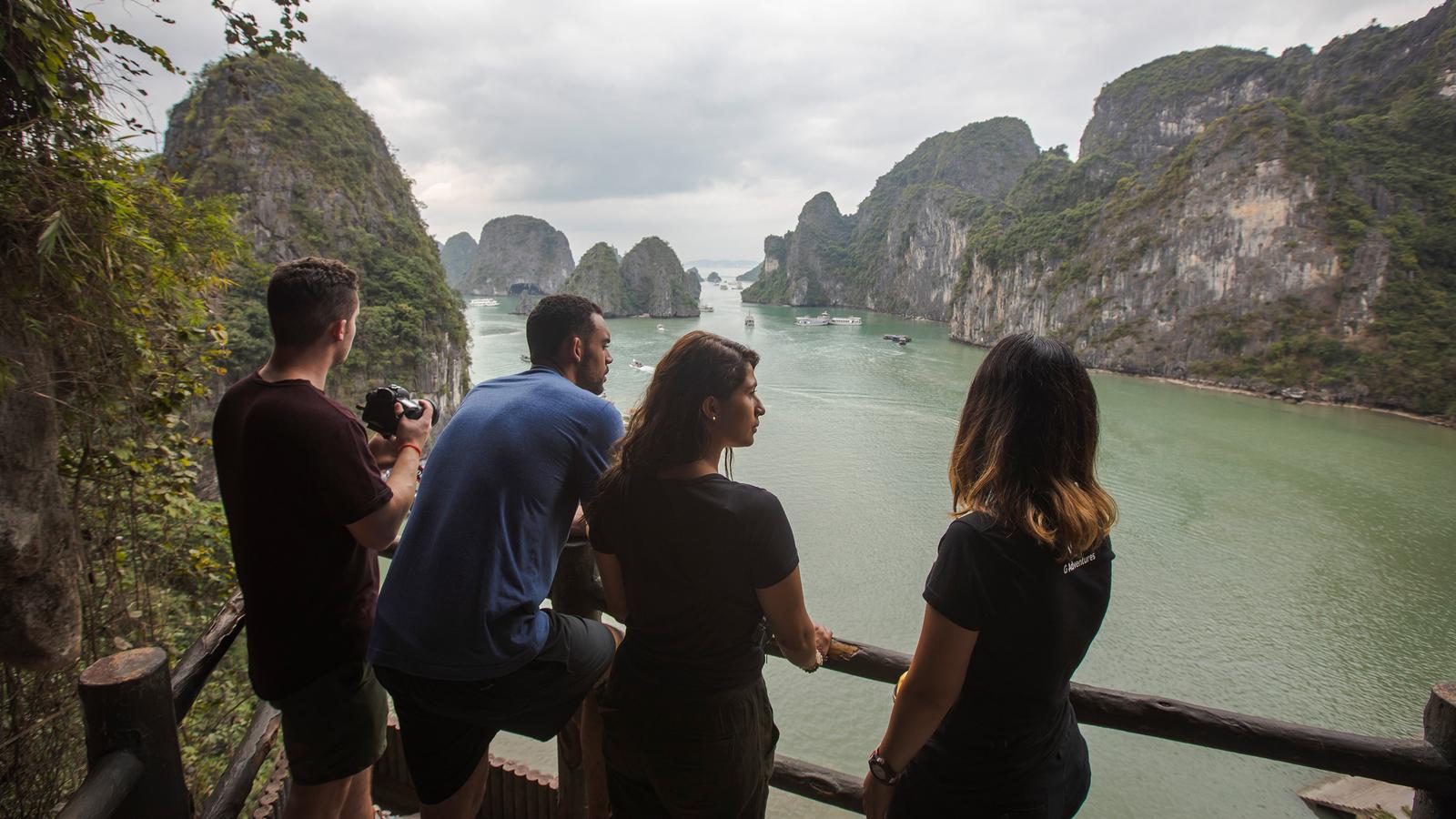A group of people standing at a wooden railed viewing platform overlooking the placid waters and towering limestone peaks of Halong Bay in Vietnam