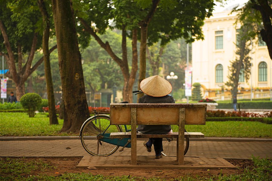 A woman in a traditional conical hat sits on a park bench in Hanoi, Vietnam