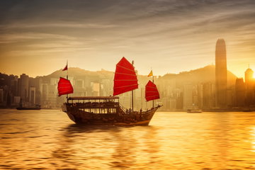 A red sailed junk boat cuts a silhouetted figure in the waters in front of Hong Kong island at sunset