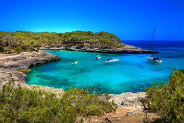 Crystal clear turquoise and azure waters, topped with small white boats floating near the rocky shores topped with green trees, in the Balearic Islands, Spain