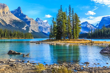 Snow-capped, jagged mountains surround the landscape of Jasper National Park, Canada, with striking slim trees and a crystal clear lake at their foot.