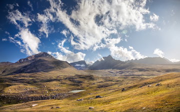 Brown-green grassed hills transform into the jagged snow-capped peaks of the mountains Kyrgyzstan rising high into a blue clouded sky