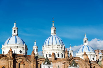 Three ornate, blue and white domes of a cathedral in Cuenca sit on sandstone arches against bright blue clear sky, Ecuador
