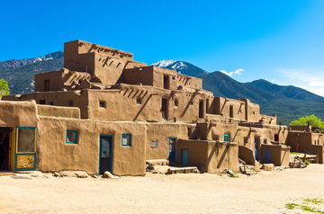 A light brown pueblo style building sits in bright sunshine, with a snow-capped forested mountain stretching out behind it, New Mexico, USA