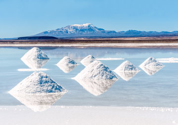 Small triangular mounds of salt piled on the reflective Salar de Uyuni with a mountain in the distance set against the blue sky