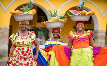 Three colourfully dressed Palenqueras (fruit sellers) women balancing bowls of exotic fruit on their heads and smiling at the camera, Cartagena, Colombia