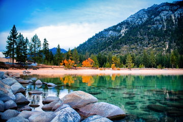 Tall mountains dusted in snow tower over an emerald green lake that laps up to a small sandy shoreline, lined with orange and green trees in Lake Tahoe, California, USA