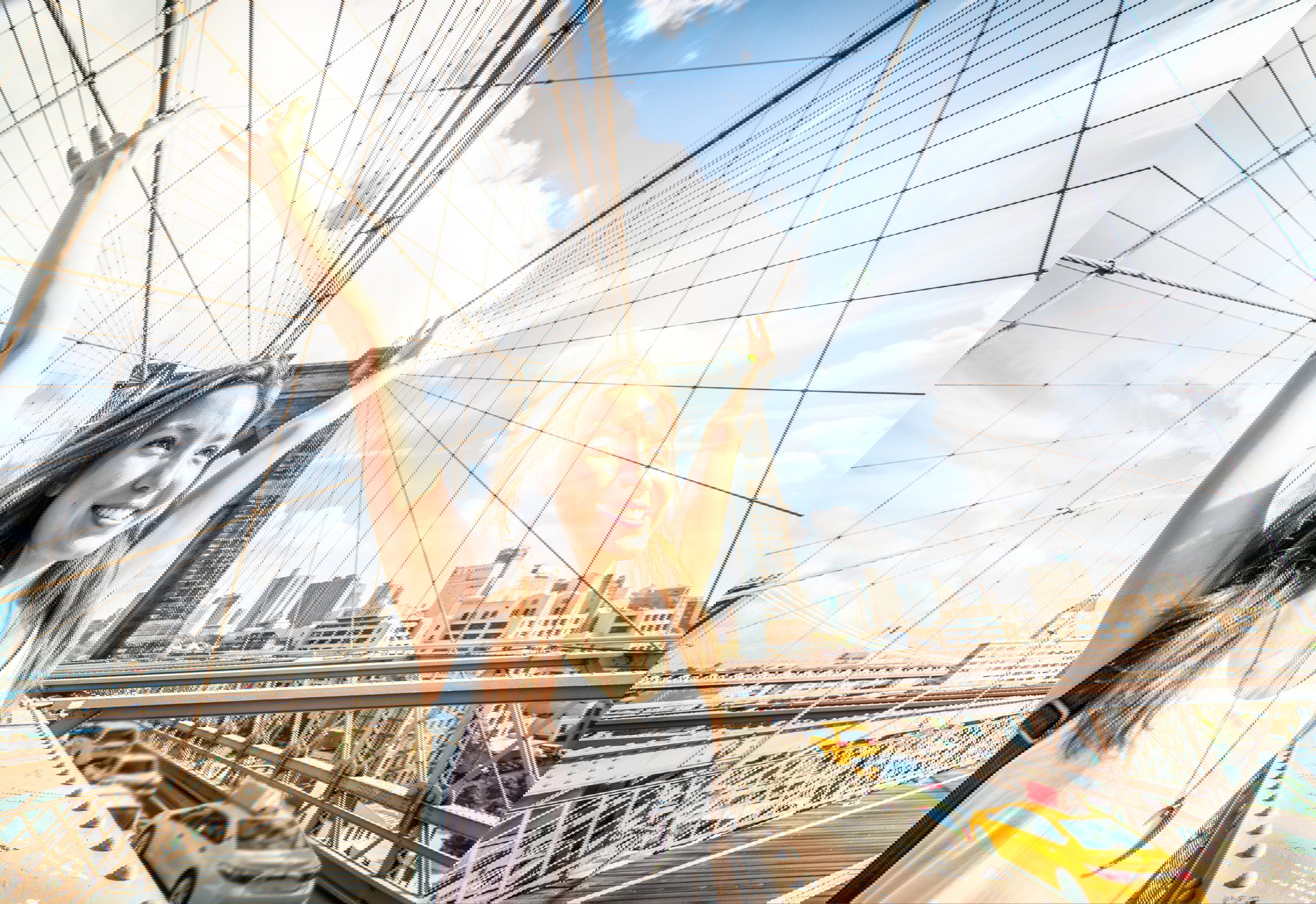 A female tourist stands on the walkway of the Brooklyn Bridge with her arms in the air and yellow cabs in the background, New York City, USA