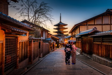 Two geishas in traditional Japanese kimonos in front of a temple at dusk in Kyoto, Japan