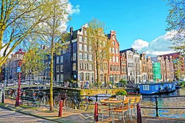 A tiny table topped with a basket of flowers next to bicycles lined up against the railings over-looking the canal with a blue house boat and the iconic tall and colourful buildings of Amsterdam, the Netherlands.
