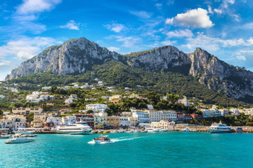 A beautiful turquoise sea fills the Bay of Capri, colourful buildings line the shore, mountains rise high in to sky behind, Italy