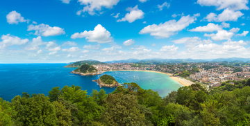 Aerial view of thick greenery giving way to views of turquoise sea near the golden sands of the beach and towns splayed across the terrain and the mountains appearing on the horizon in San Sebastian, Spain