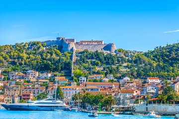 A sleek white ship sits at the dock in front of a red roofed buildings dot a hill covered with trees, Hvar, Croatia