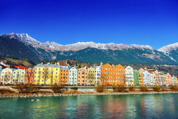 A row of tall, colourful buildings line the water’s edge in front of enormous snow-capped mountains in Innsbruck, Austria