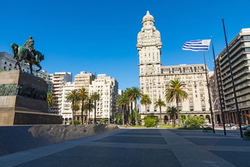 A monument of a man on a horse stands opposite the grand Palacio Salvo building with the Uruguay flag on a pole, Montevideo, Uruguay