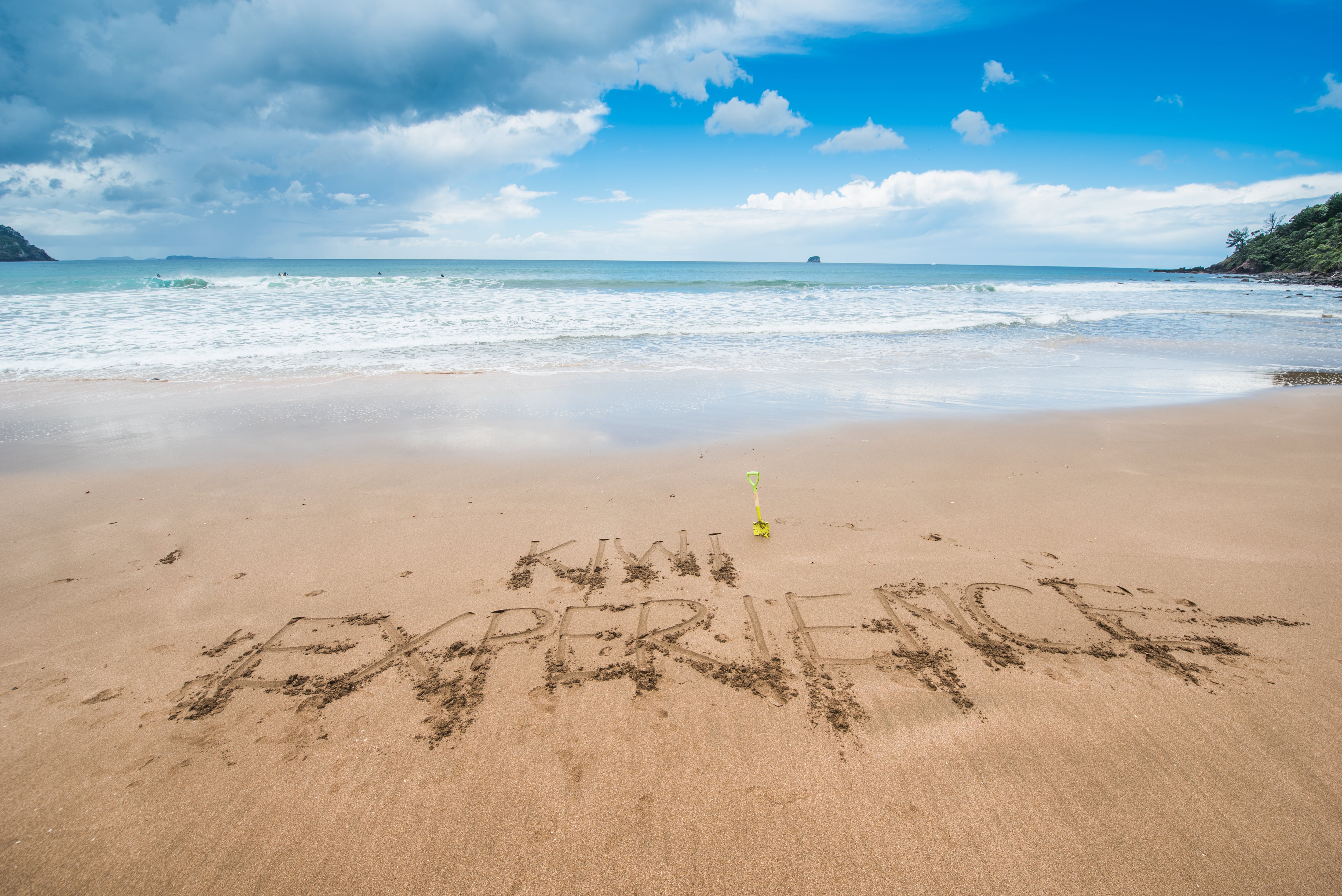 Kiwi Experience has been written into the sand, as the calm waves hit the shore on a bright day in New Zealand