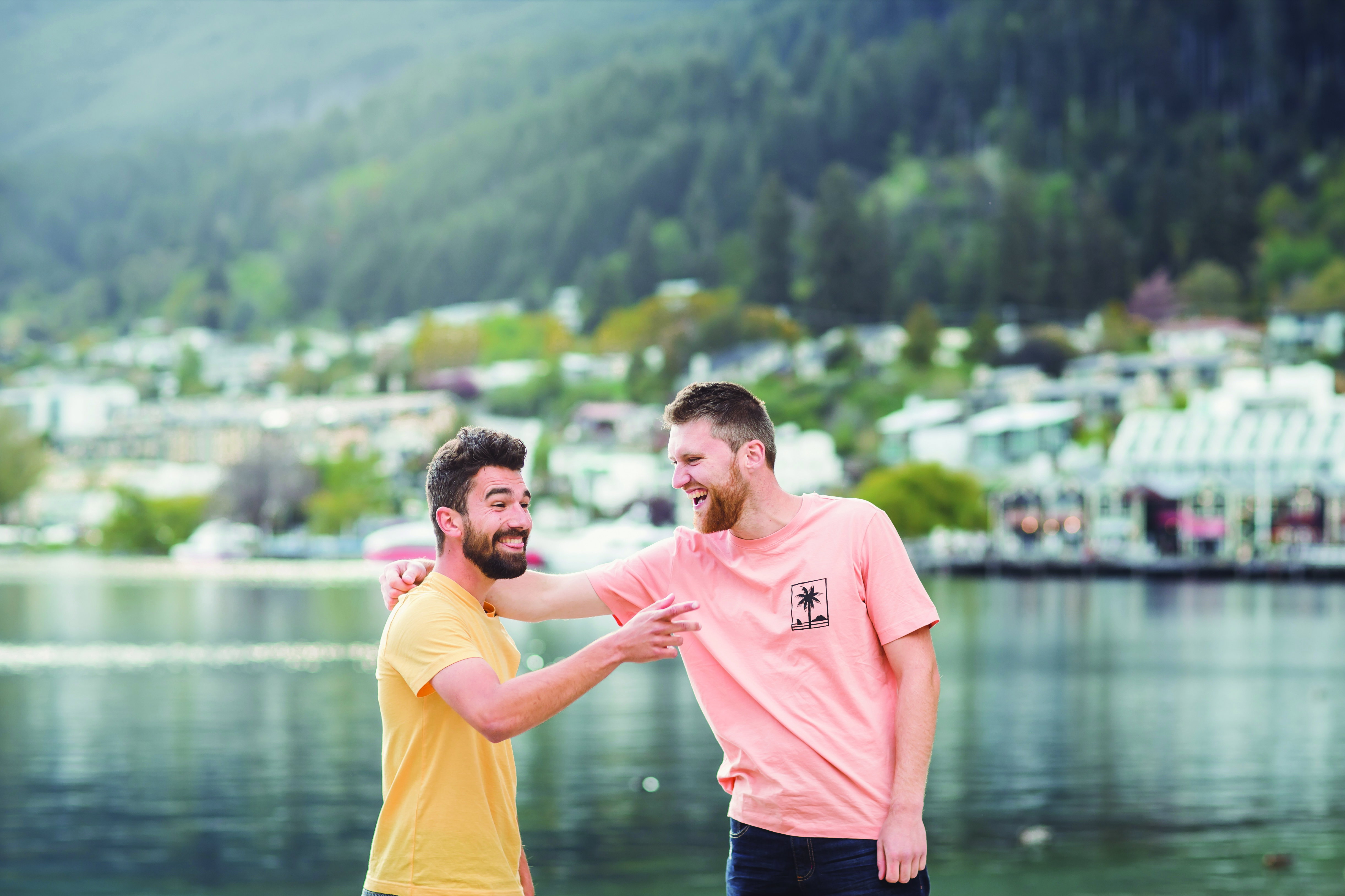 Two young men laugh and hug, whilst stood in front of a large lake, on a cloudy day in New Zealand