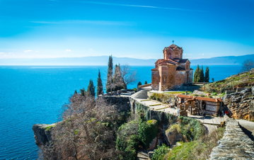 A number of steps leading towards a beautiful brick church perched on the edge of Lake Ohrid, bordered with tall trees, North Macedonia
