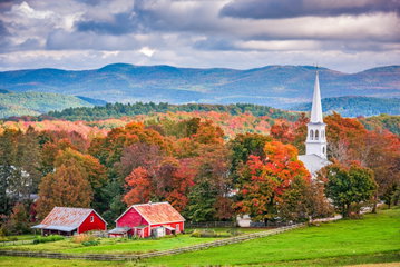A red farm house is sheltered by a lush forest; full of trees of all shades of reds, oranges and greens, showing how beautiful the autumn is across New England in the USA