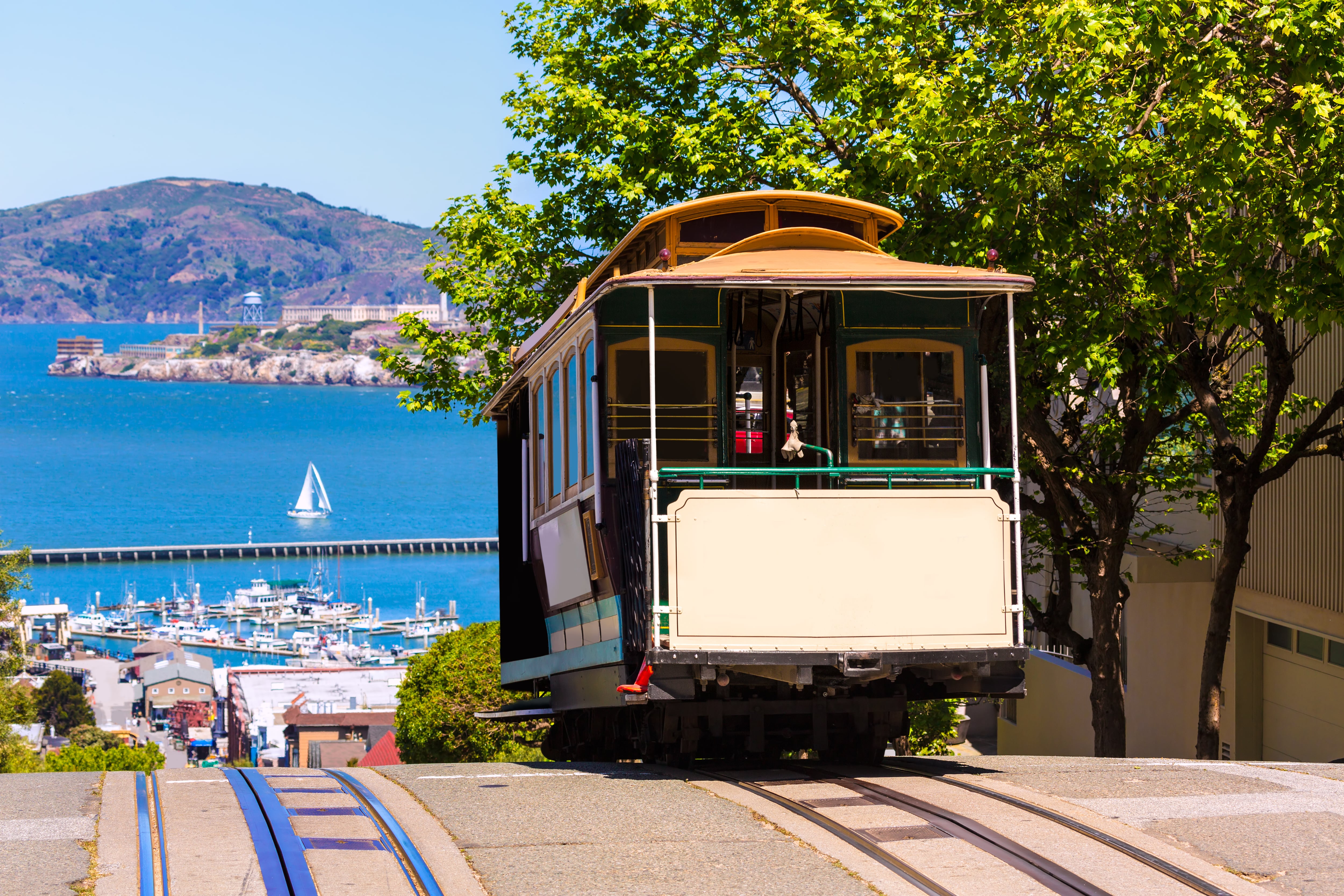 a historic cable car is at the crest of a steep hill in San Francisco and in the background is the blue water of the bay and sat within it is the infamous Alcatraz prison