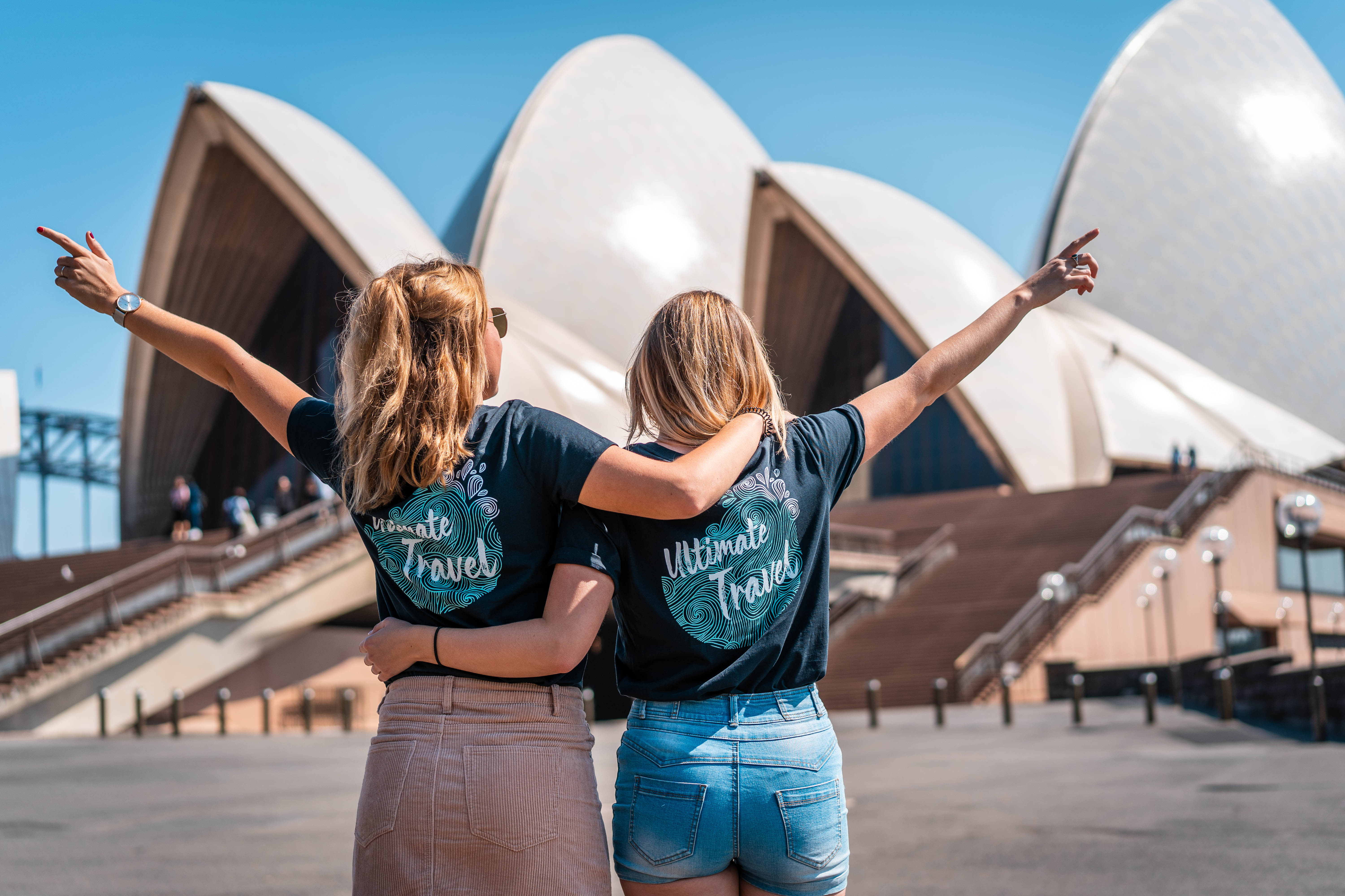Two blonde young women wearing black Ultimate branded t-shirts face the Sydney Opera House with one arm around each other and the other raised in the air with excitement.