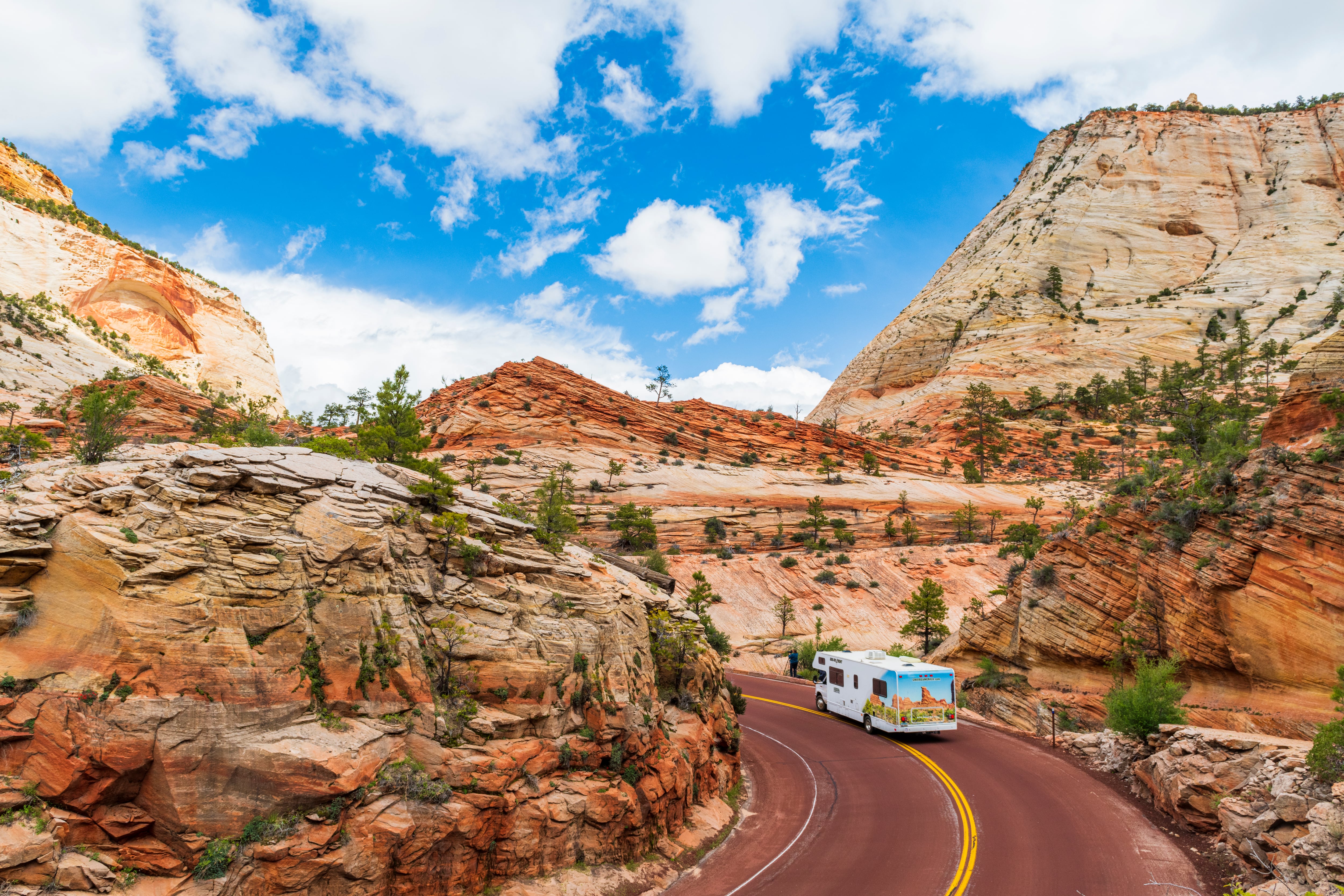 A Cruise America motorhome driving through the rugged cliffs of Zion National Park with a bright blue, cloudy sky above Utah, USA