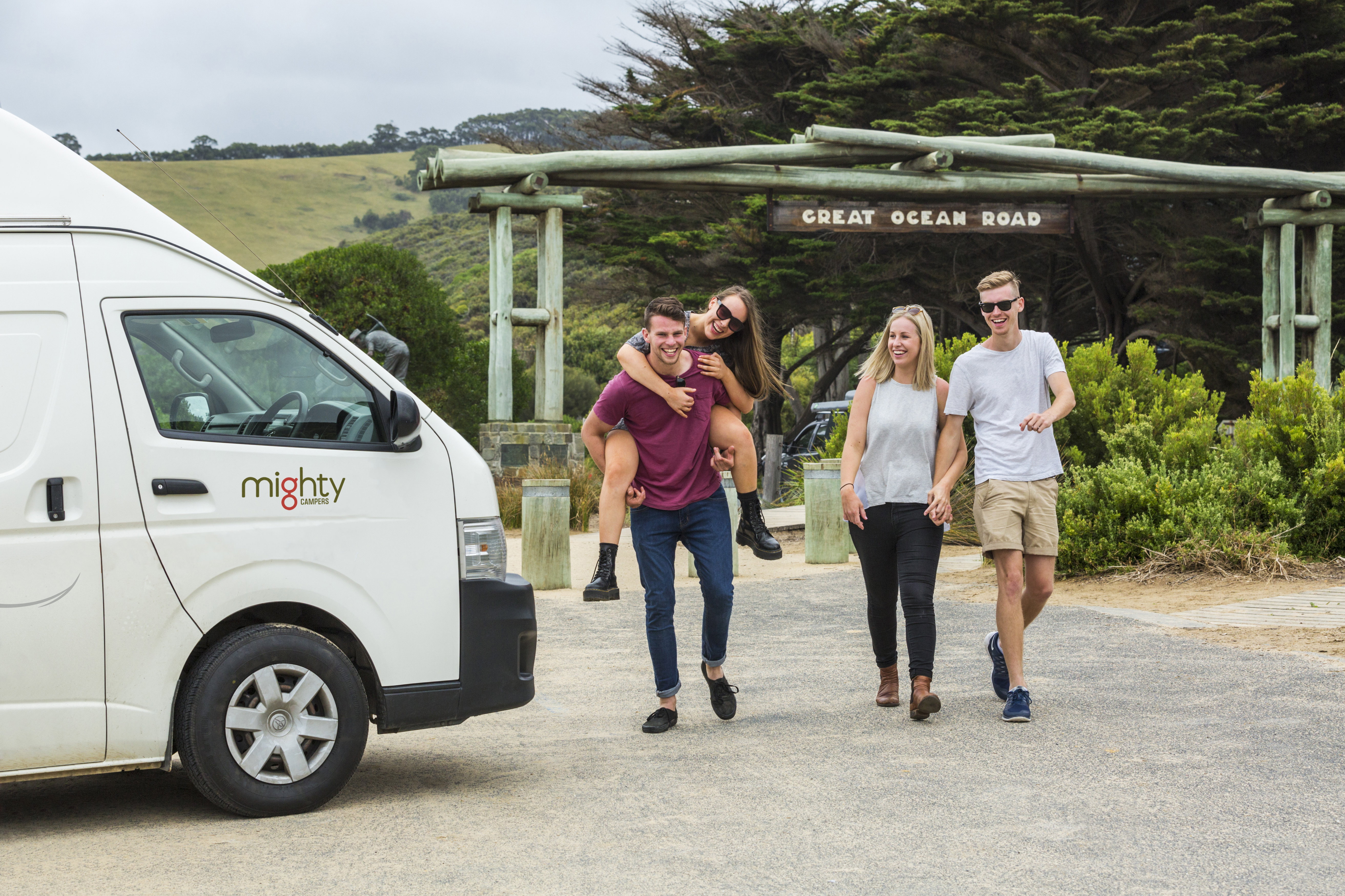A couple holding hands and a woman piggy backing on a friend in front of a Mighty Camper and the Great Ocean Road Sign, Victoria, Australia
