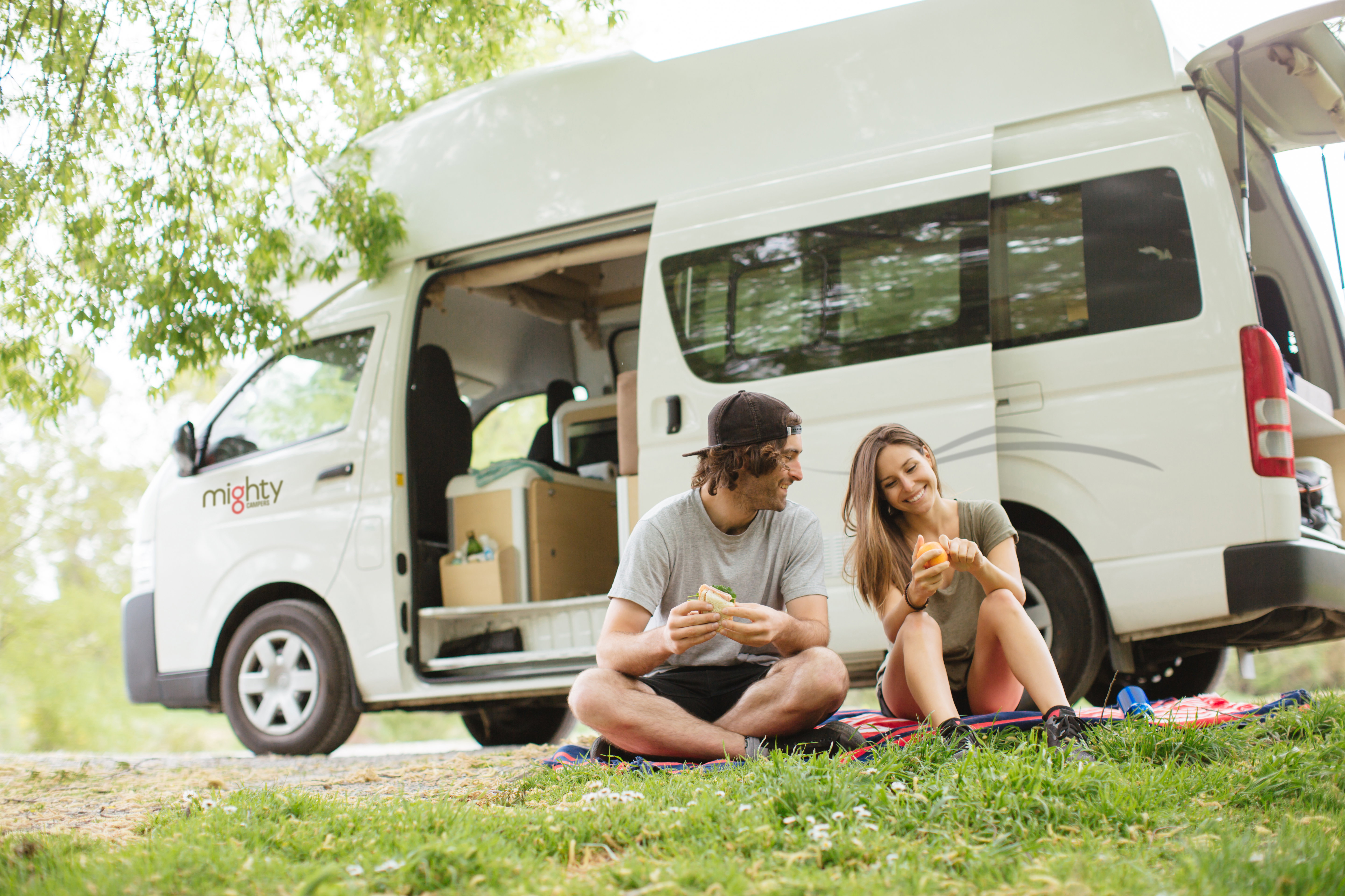 A couple sat in front of a Mighty Campervan eating a sandwich and an orange on a picnic blanket just off the road in New Zealand