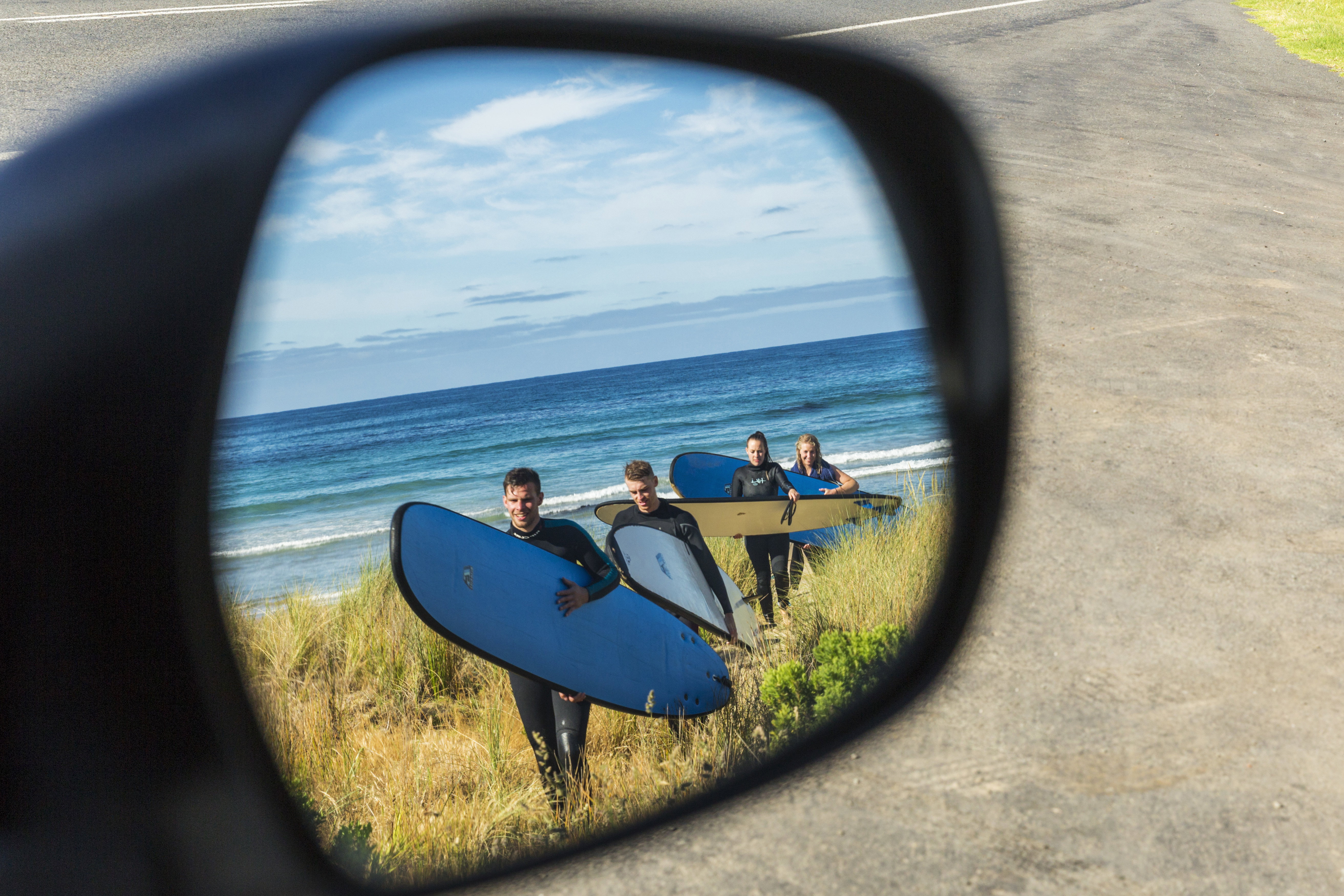 Two couples wearing wet suits walk from the beach clutching their surf boards as they make their way back to their Mighty Campervan, as seen from the perspective of the side rear view mirror, New Zealand
