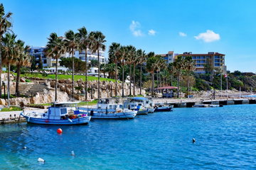 Blue sea glinting vibrant in the sun, with fishing boats hugging the promenade lined with tall palm trees blowing in the wind, the grass behind them leading up to the buildings behind, Paphos, Cyprus