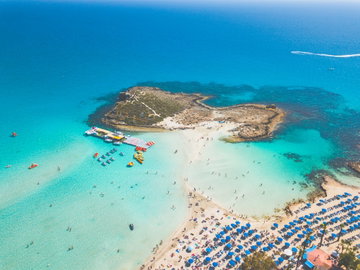 Aerial view of a pure white sand beach, filled with blue umbrellas and people swimming in the crystal clear sea and a strip of white sand leading to a small archipelago in Larnaca, Cyprus