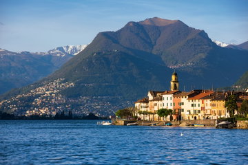 Orange, yellow and white houses on shore of Lake Lugano with rolling hills and snow-capped mountain in the background, Switzerland