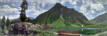 A jagged, rocky peak bursts from the lush green surrounding hillside behind a glassy lake and a red roofed building, Pakistan