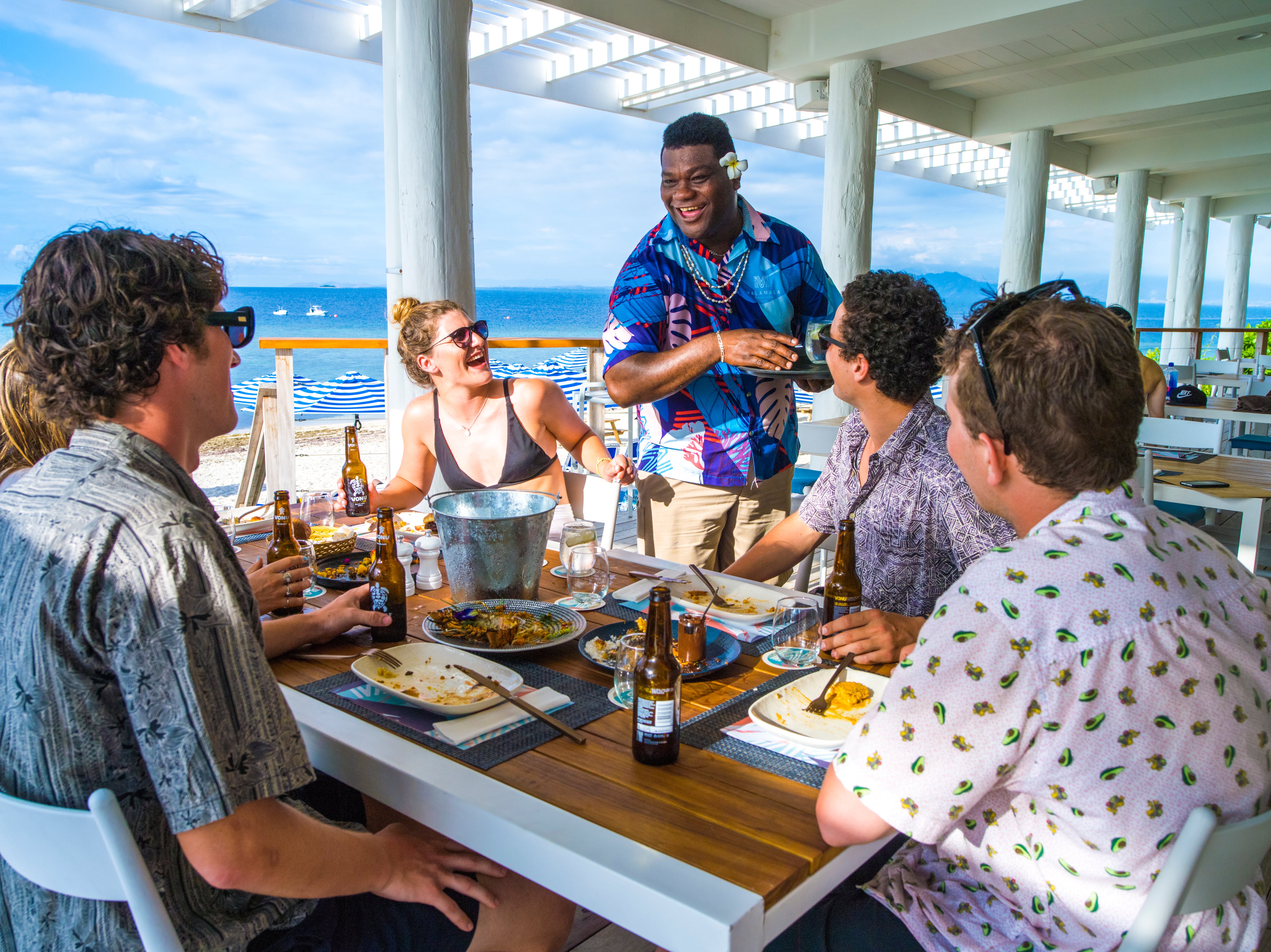A Fijian waiter in a colourful shirt laughs with tourists who are sat at a table, enjoying a meal by the ocean.