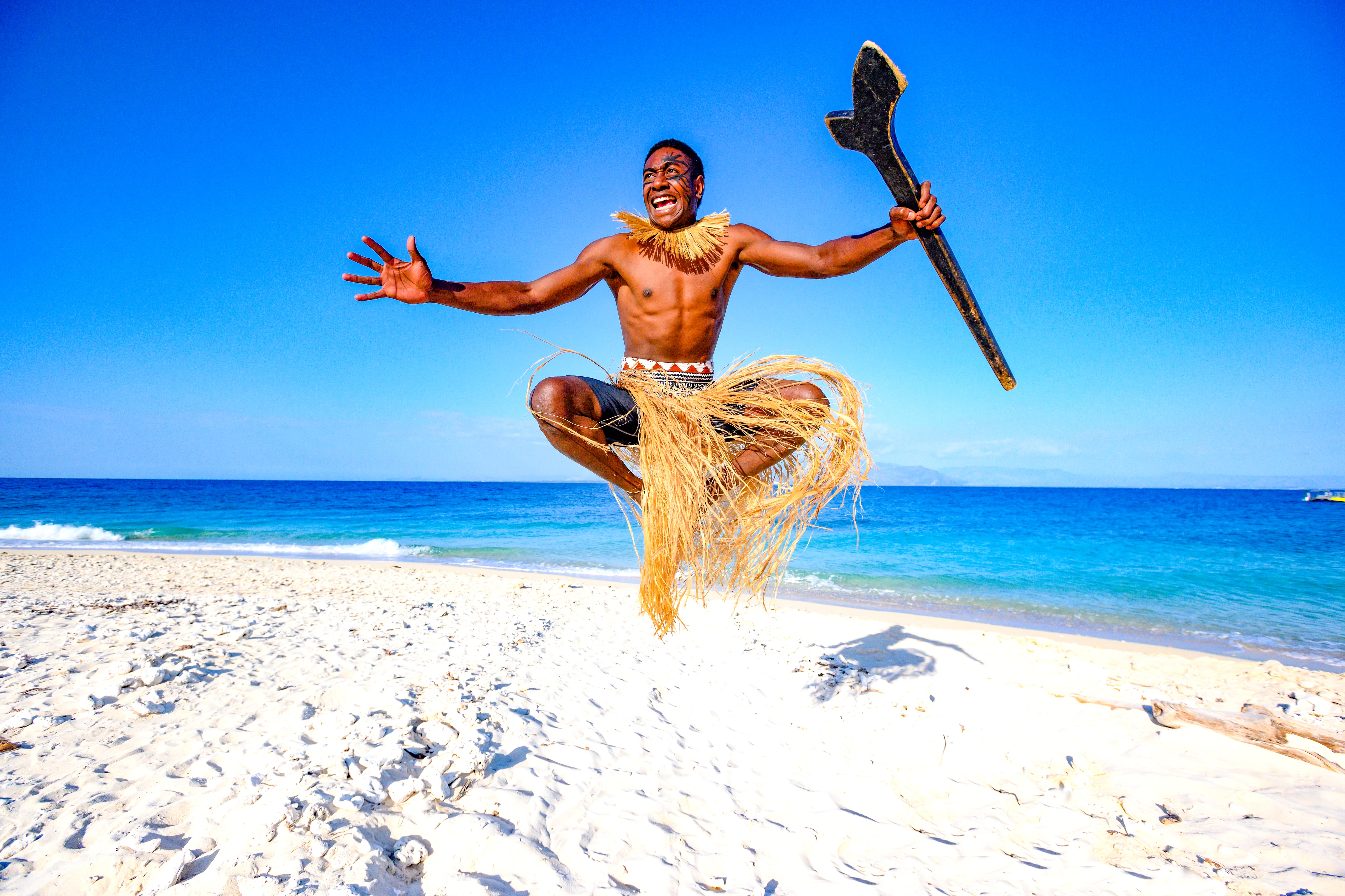 A smiling male Fijian Kuata warrior dressed in a grass skirt jumps into the air on a white sand beach