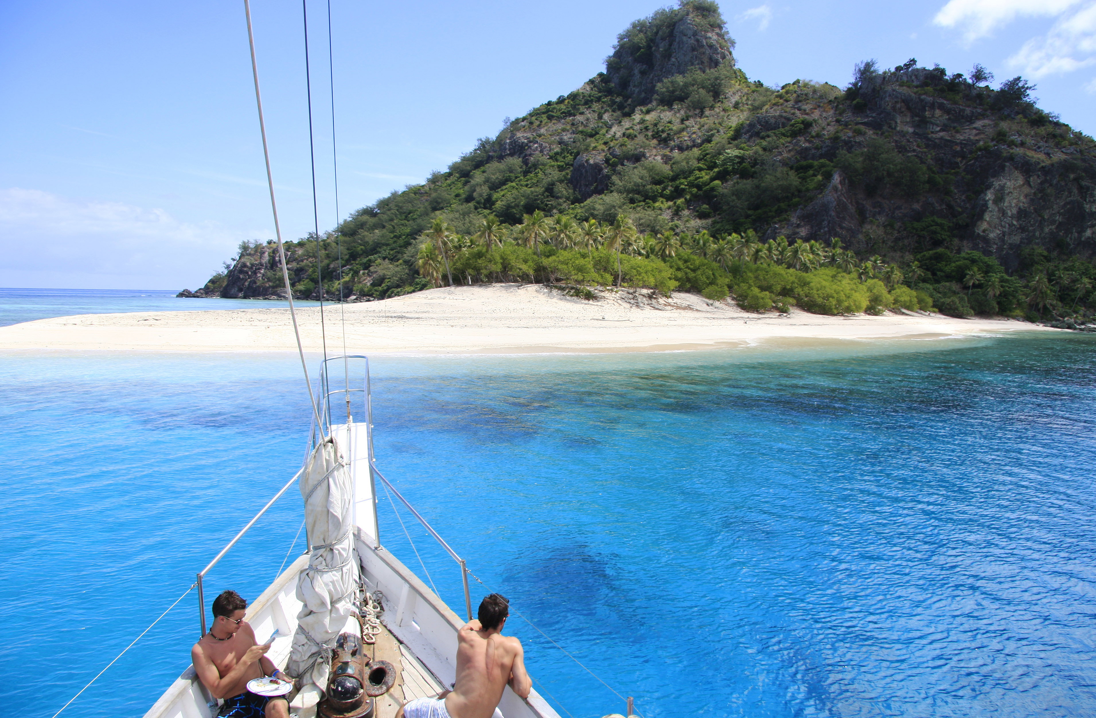 The front of a sailboat approaches a Fijian tropical island, with white sand beach against a jungle clad mountainous background.