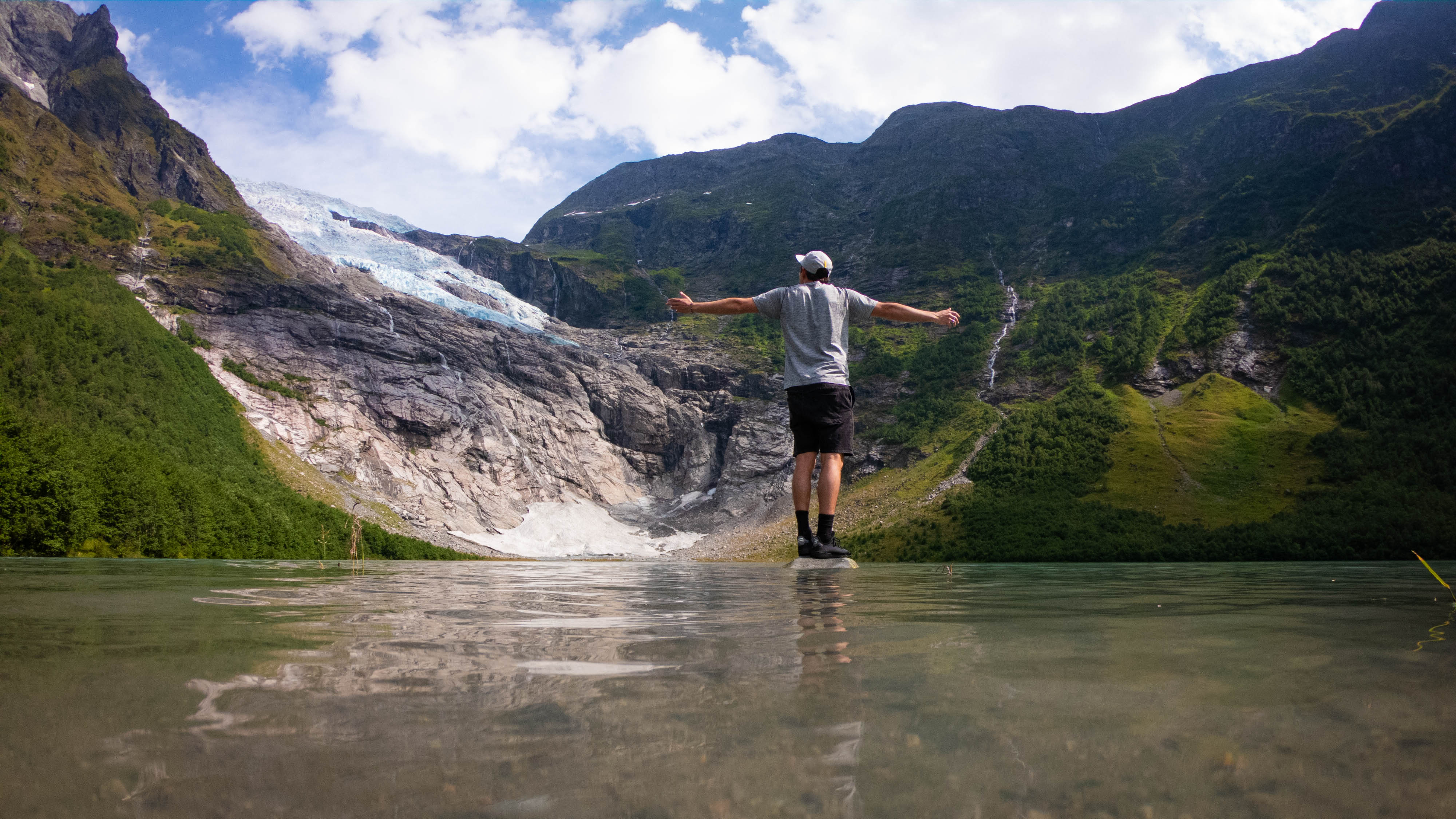 A young male in a cap and a grey t shirt stands on a rock in the middle of a river with arms outstretched, facing dramatic fjord scenery.