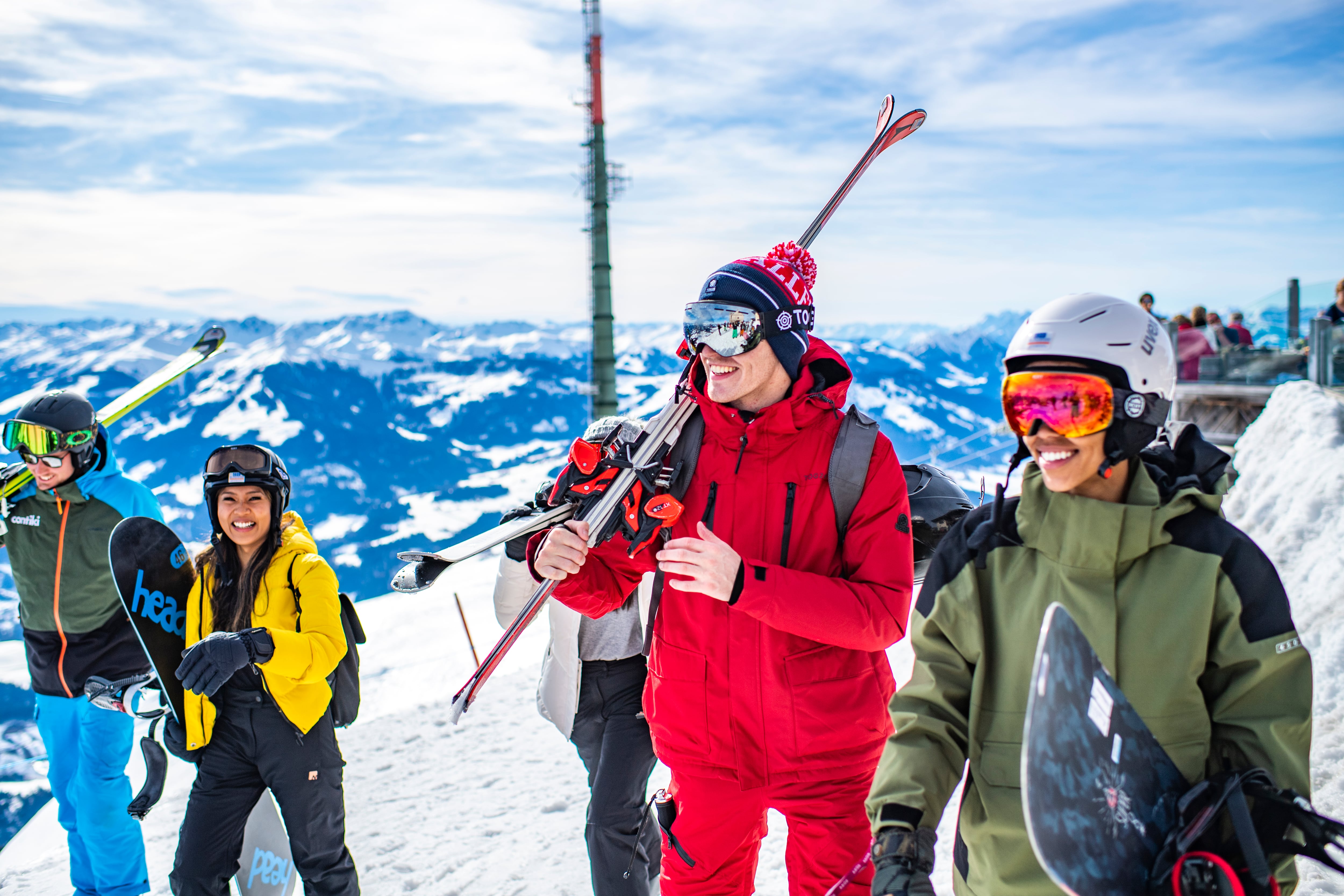 A group of young smiling travellers carry their skis and snowboards across snowy slopes against a backdrop of mountain scenery.