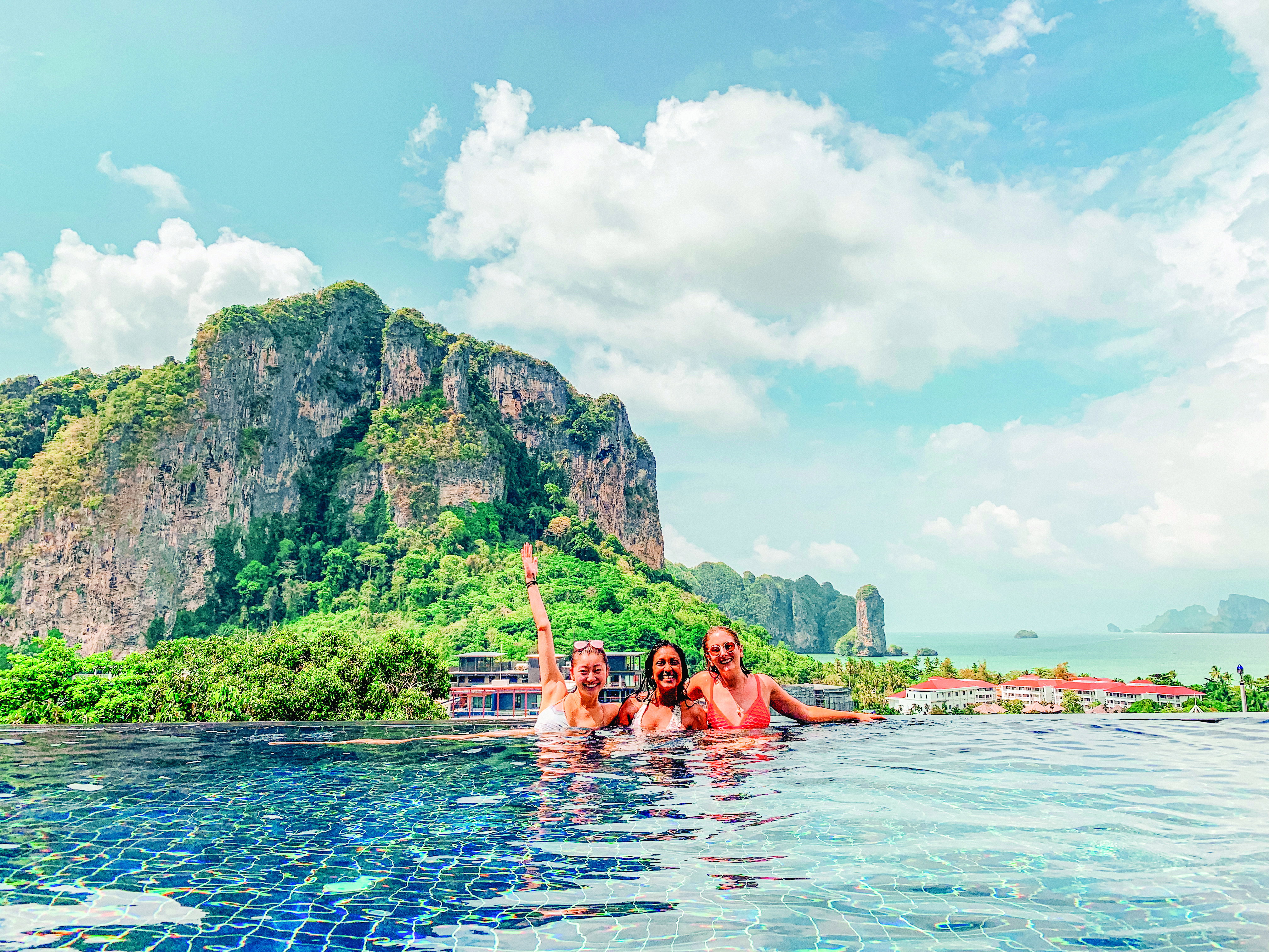 Three young women half submerged in a swimming pool smile, with jungle clad limestone carst mountains behind them.