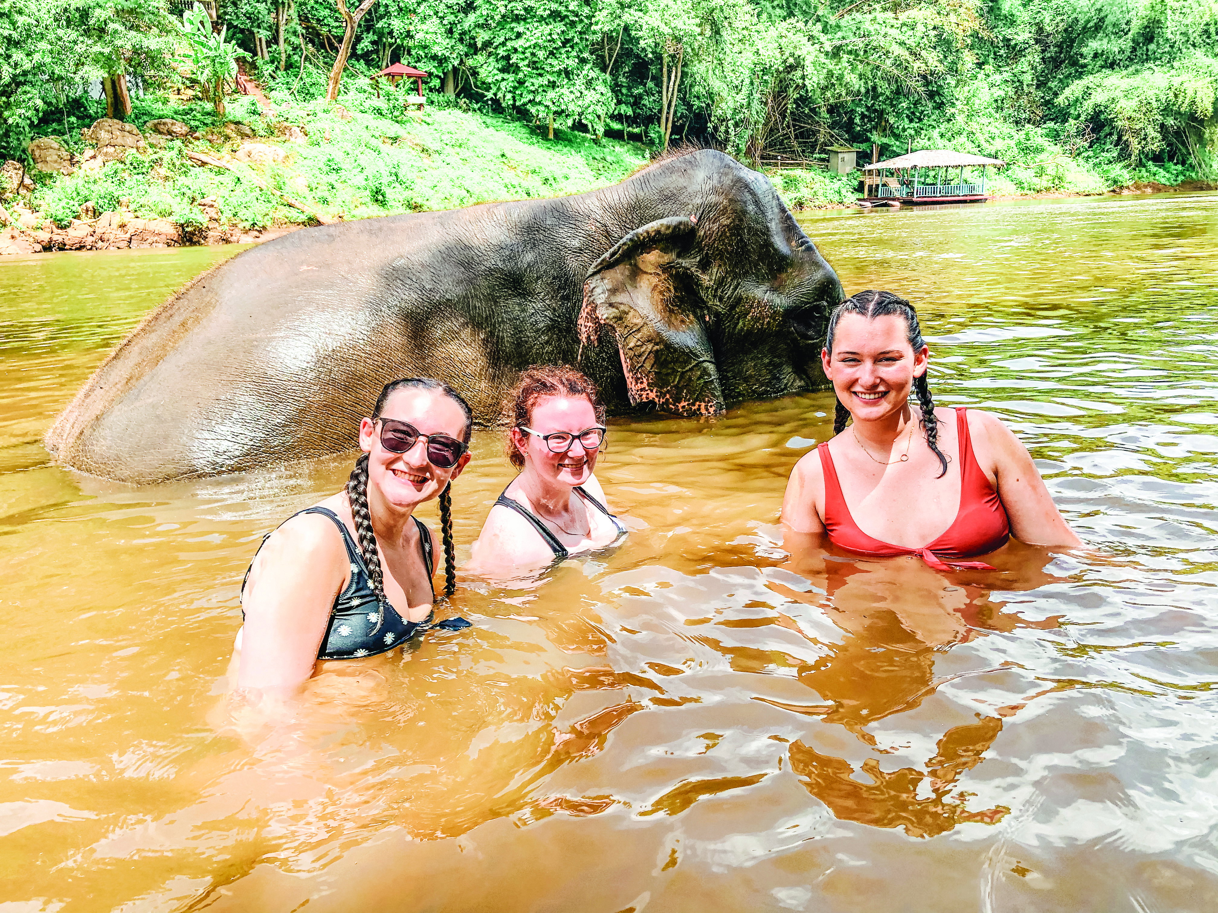 Three girls are half submerged in brown river waters with an elephant bathing behind them.