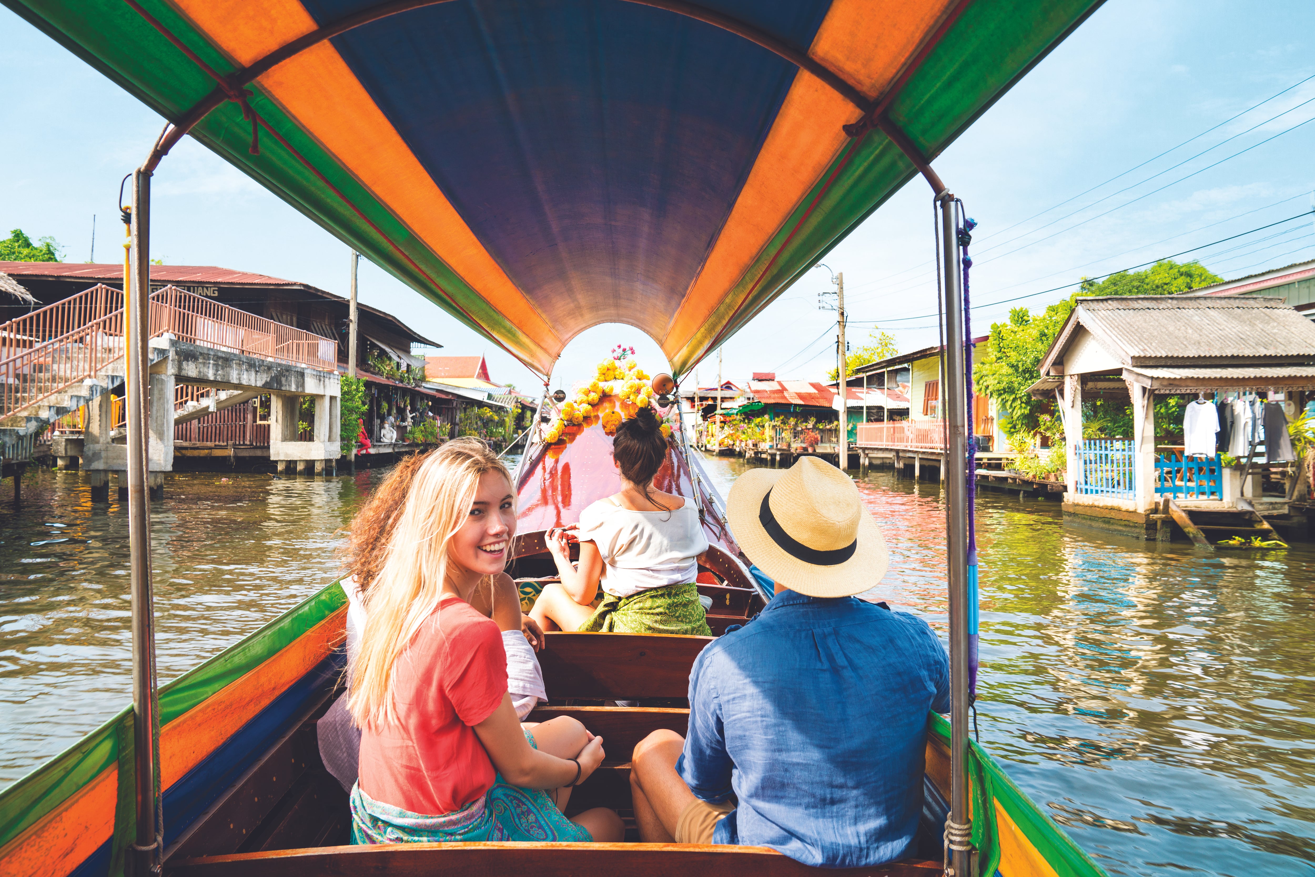 Three travellers sit in a water taxi heading down the river in Bangkok, with one girl looking back towards the camera, smiling,