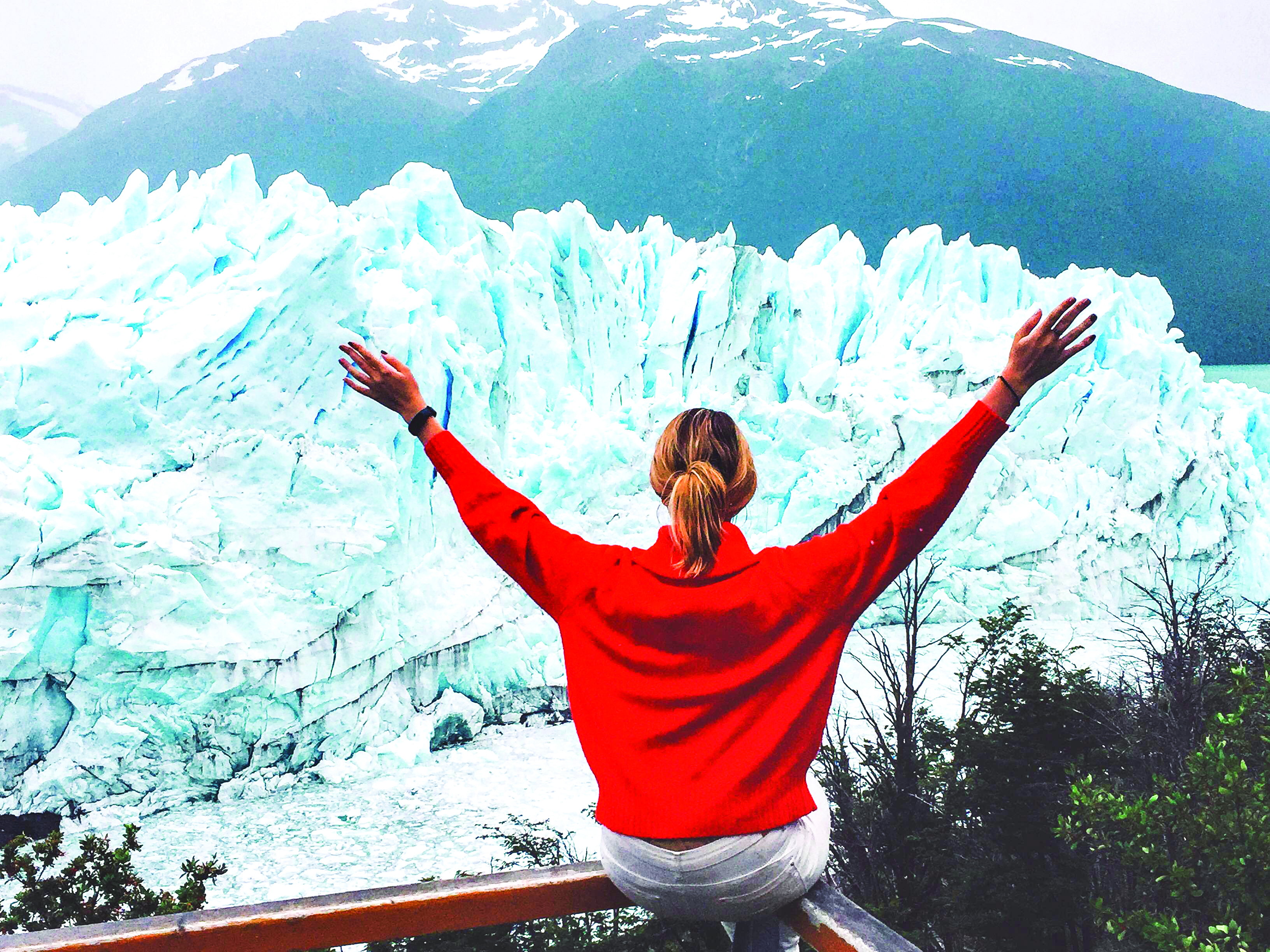A female in a red jumper raises her arms in the air as she faces the jagged ice walll of the perito merino glacier. 