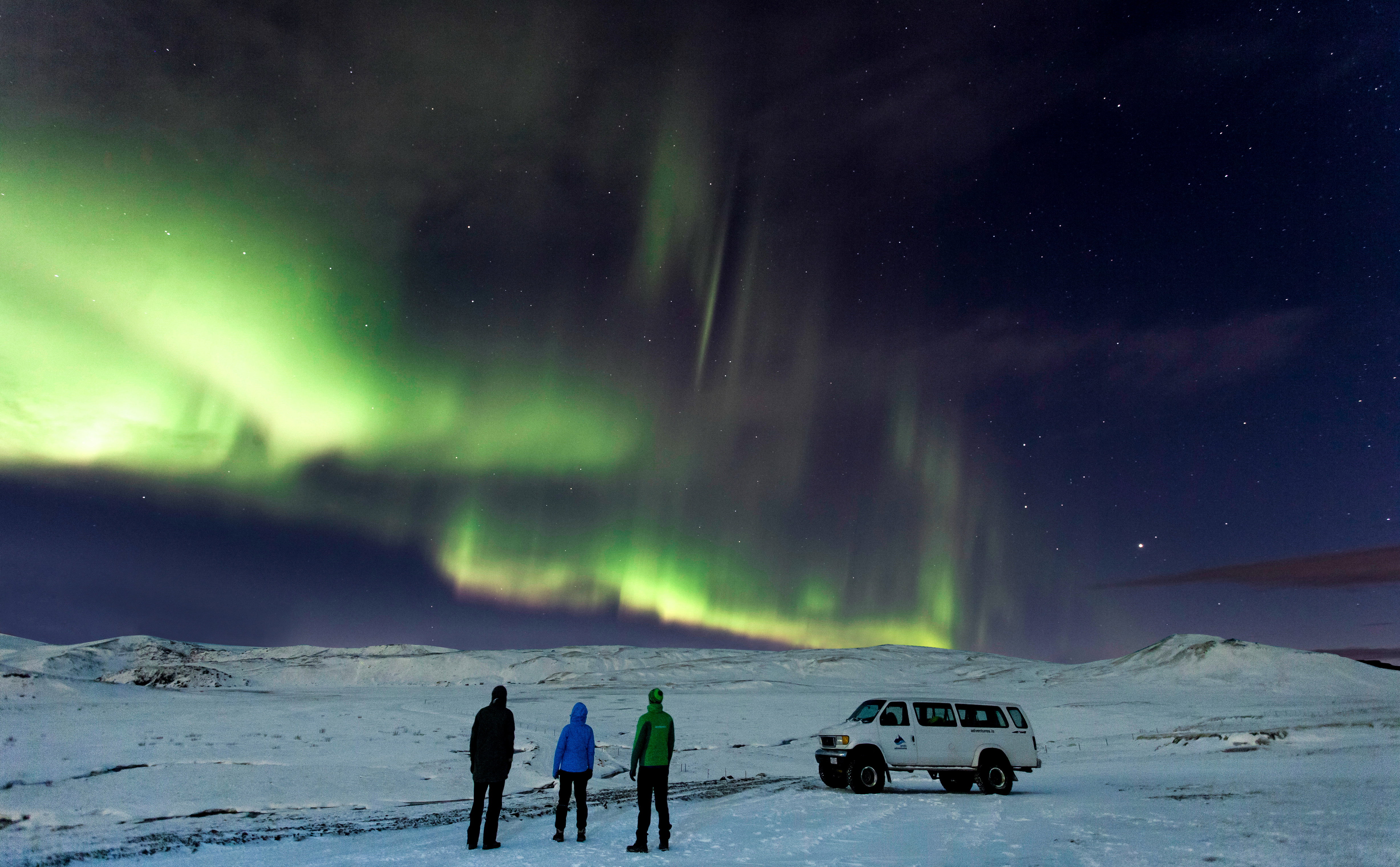 Three people stand next to their 4WD in the snowy mountains of Iceland as the green and orange Northern Lights dance in the sky.