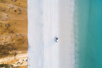 An aerial view of a jeep driving along a white sand beach flanked by orange cliffs on one side and blue sea to the other, Fraser Island, Queensland