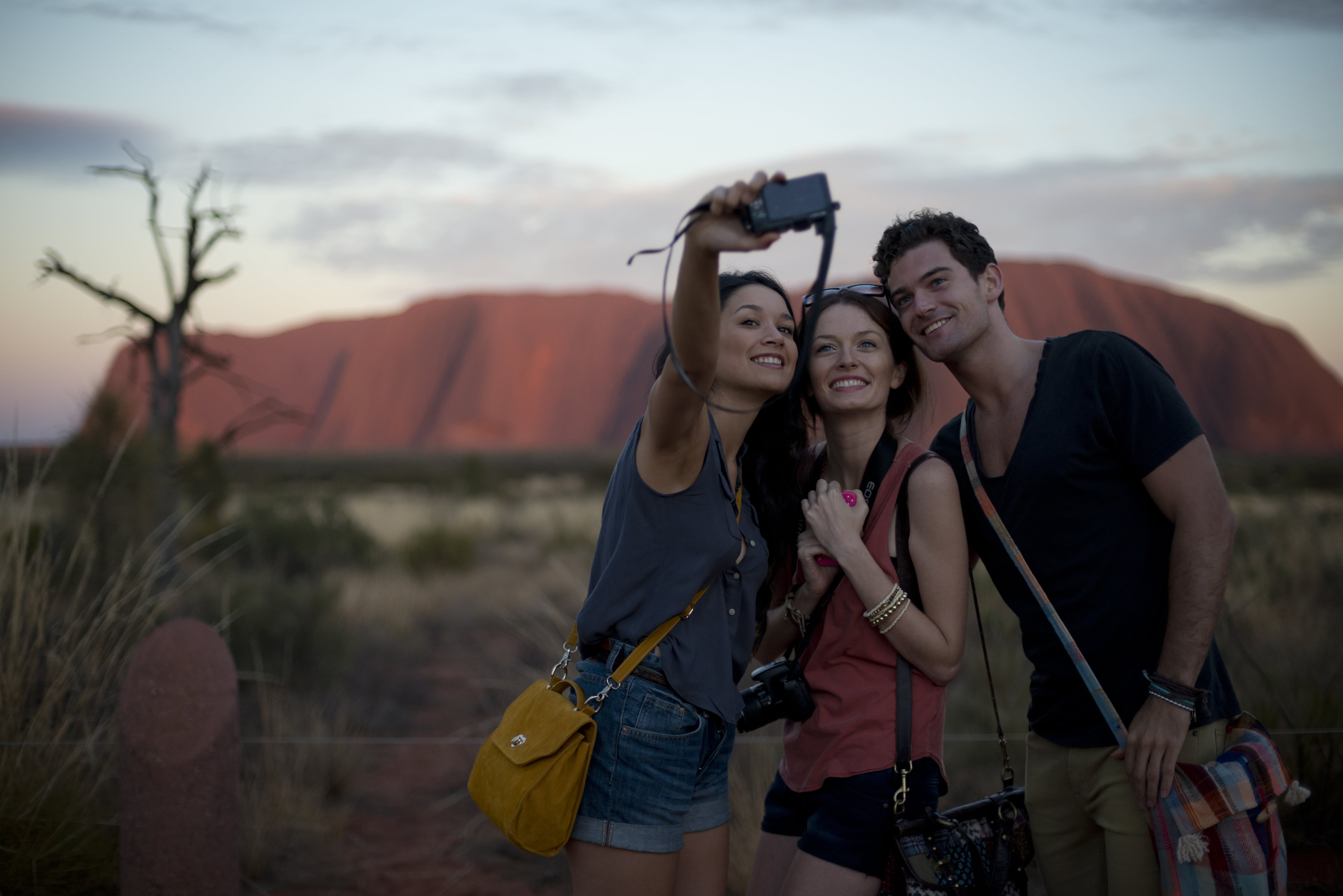 Two young women and a young man smile as they take a selfie together in front of the red, rocky escarpment of Uluru. 