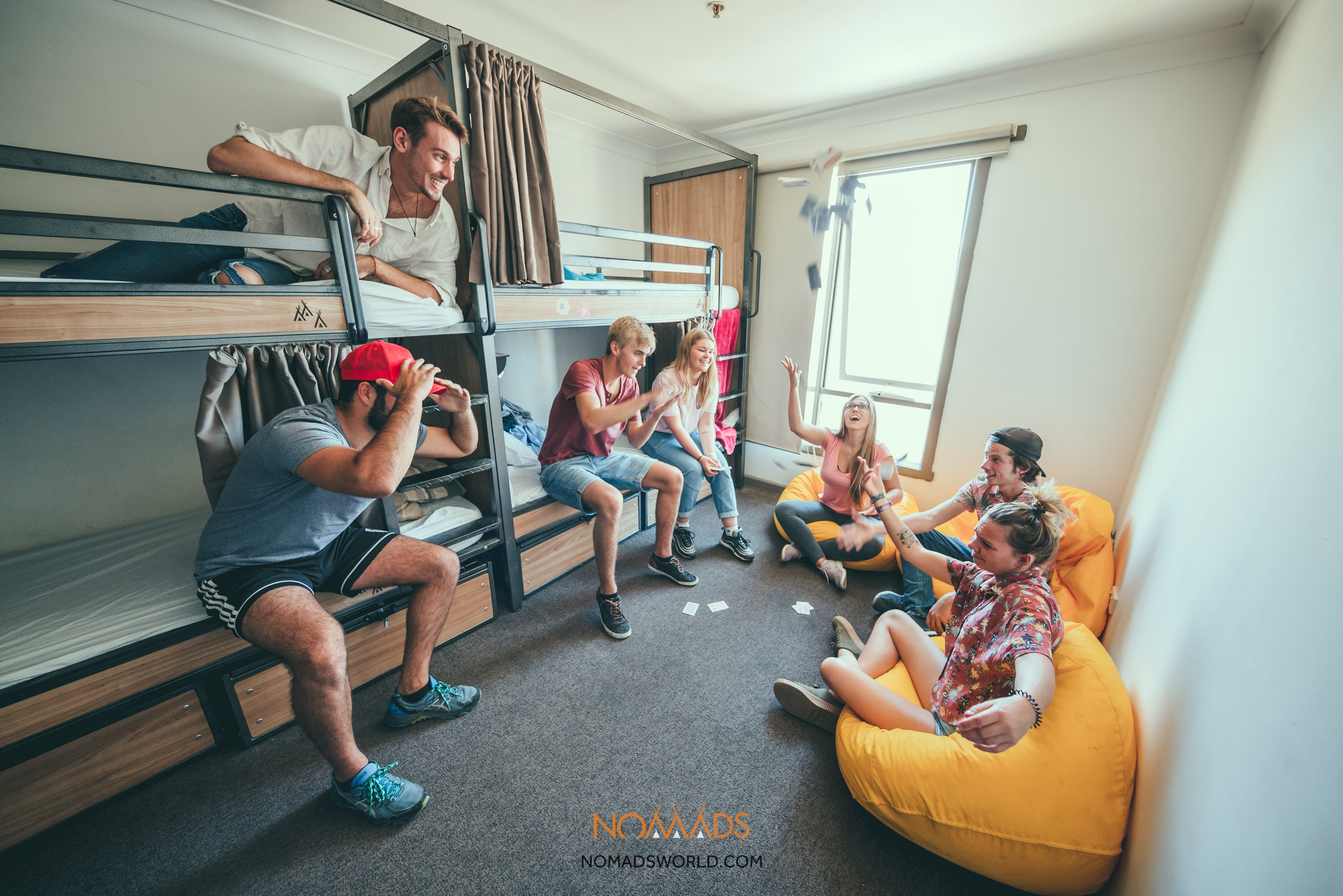 A group of seven backpackers hang out in a hostel dorm, sat on bunk beds and yellow bean bags, exclaiming as one girl throws a deck of cards in the air. 