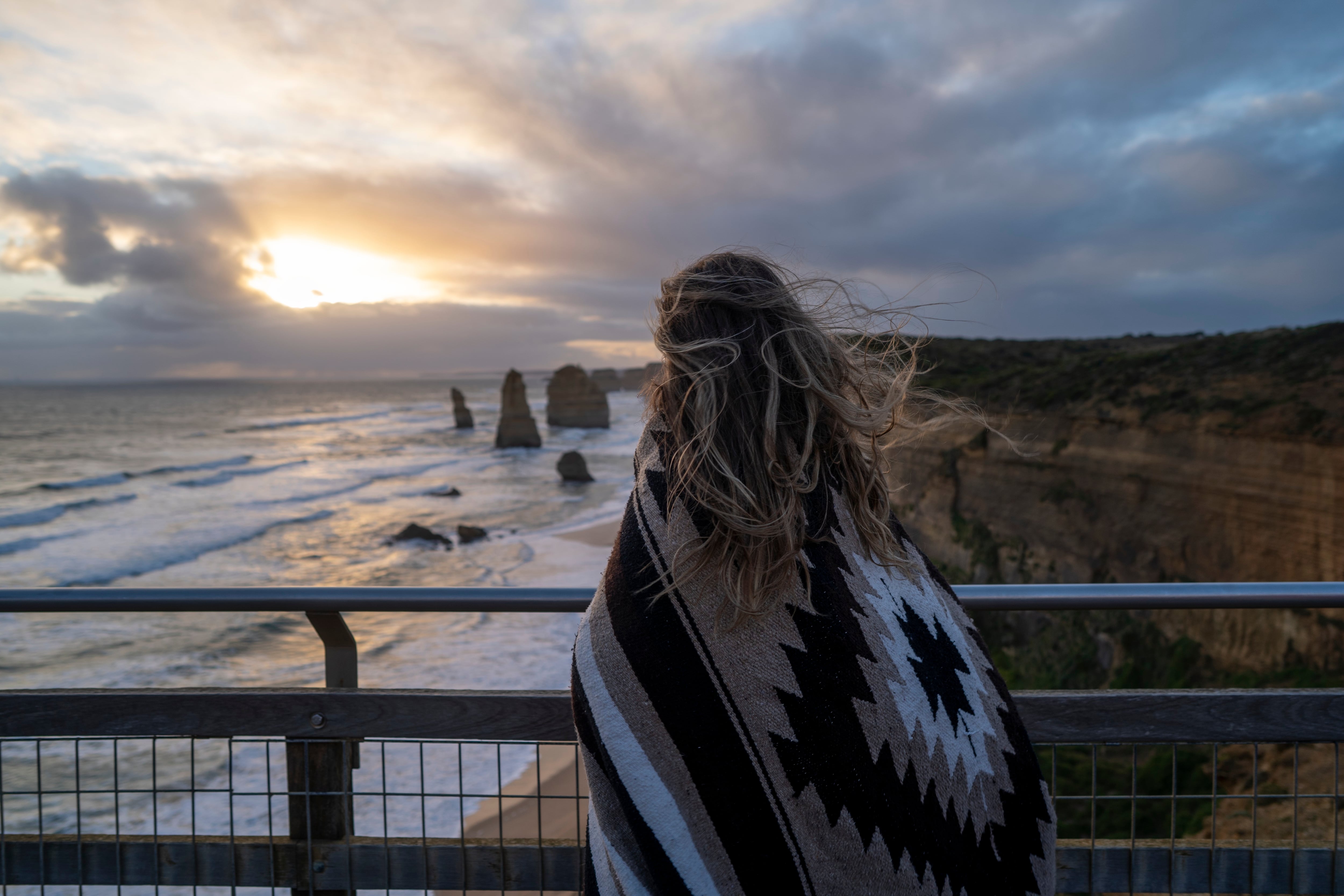 A young woman holds a patterned blanket round her shoulders as she watches the sunset over the Twelve Apostles, waves lapping at the beach and her hair blowing in the breeze.