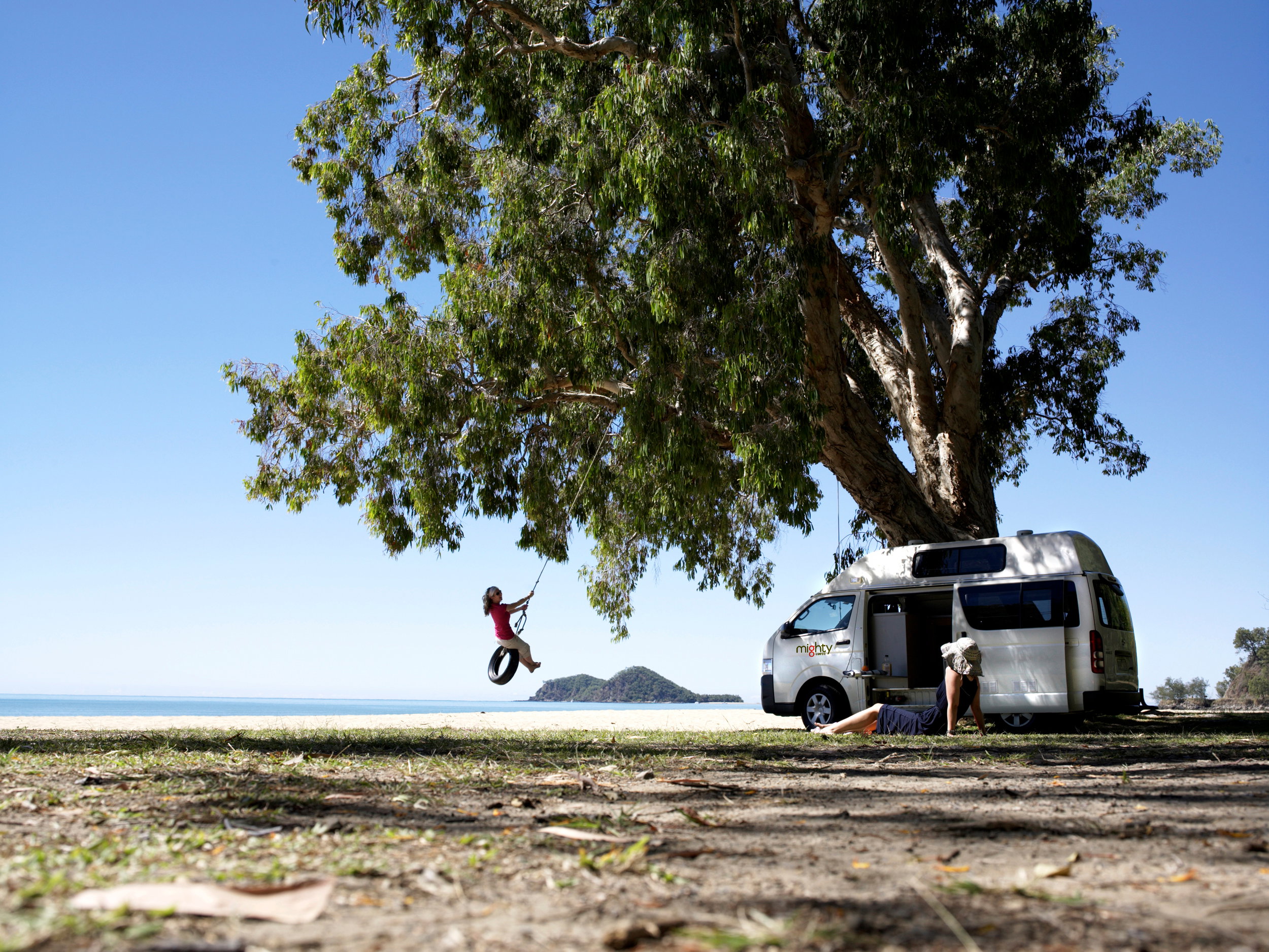 A woman swinging on a tyre swing from a tree on the beach, while another woman lounges in front of their Mighty Camper, Tasmania, Australia